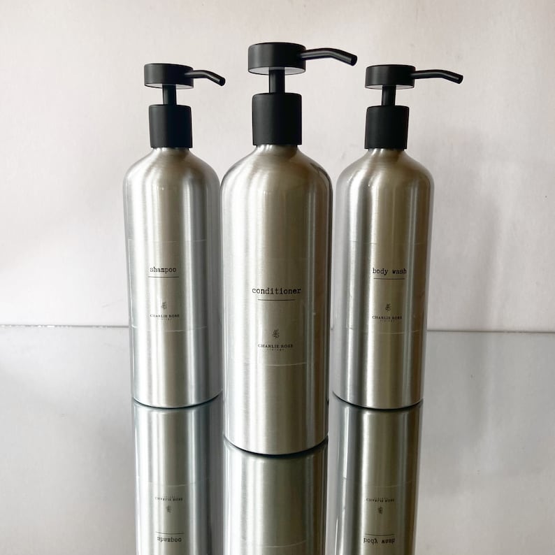 May include: Three silver pump bottles with black pumps and labels. The bottles are labelled "shampoo", "conditioner", and "body wash".
