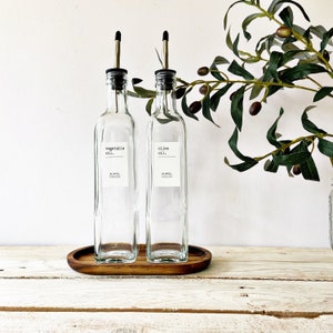 May include: Two clear glass bottles with black pour spouts and labels that read "vegetable oil." and "olive oil." The bottles are sitting on a wooden tray.