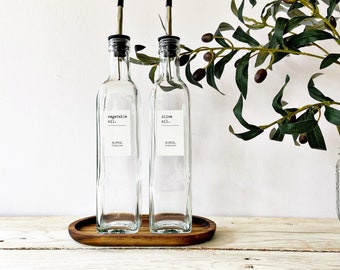 May include: Two clear glass bottles with black pour spouts and labels that read "vegetable oil." and "olive oil." The bottles are sitting on a wooden tray.