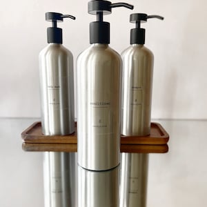 May include: Three silver bottles with black pumps and labels. The bottles are labelled "body wash", "conditioner", and "shampoo". The bottles are on a wooden tray.