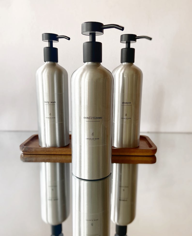 May include: Three silver bottles with black pumps and labels. The bottles are labelled "body wash", "conditioner", and "shampoo". The bottles are on a wooden tray.