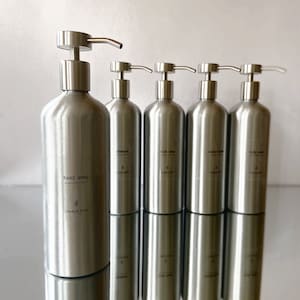 May include: Five silver pump bottles with labels. The first bottle is labelled "hand soap" with the text "CHARLIE ROSE" below. The other bottles are labelled "shampoo", "hair oil", "conditioner", and "body wash".