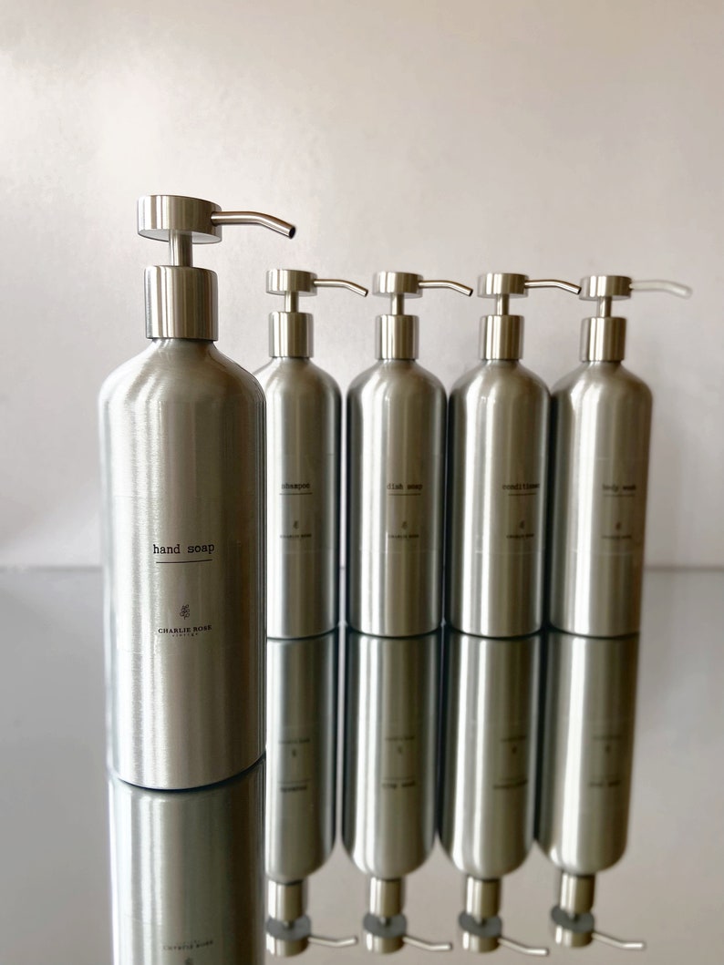 May include: Five silver pump bottles with labels. The first bottle is labelled "hand soap" with the text "CHARLIE ROSE" below. The other bottles are labelled "shampoo", "hair oil", "conditioner", and "body wash".