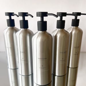 May include: Five silver pump bottles with black pumps and labels. The labels read "hand soap", "body wash", "conditioner", "shampoo", and "bath soap". Each bottle has a small floral design and the text "CHARLIE ROSE" on the label.