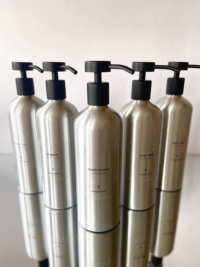 May include: Five silver pump bottles with black pumps and labels. The labels read "hand soap", "body wash", "conditioner", "shampoo", and "bath soap". Each bottle has a small floral design and the text "CHARLIE ROSE" on the label.