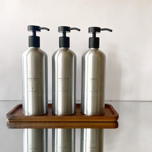 May include: Three silver bottles with black pump tops on a wooden tray. The bottles are labelled "body wash", "conditioner", and "shampoo".