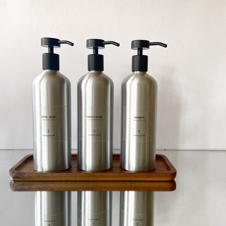 May include: Three silver bottles with black pump tops on a wooden tray. The bottles are labelled "body wash", "conditioner", and "shampoo".