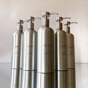 May include: Five silver pump bottles with labels for shampoo, dish soap, hand soap, conditioner, and body wash. The bottles are all the same size and shape, and they are arranged in a row on a white surface.
