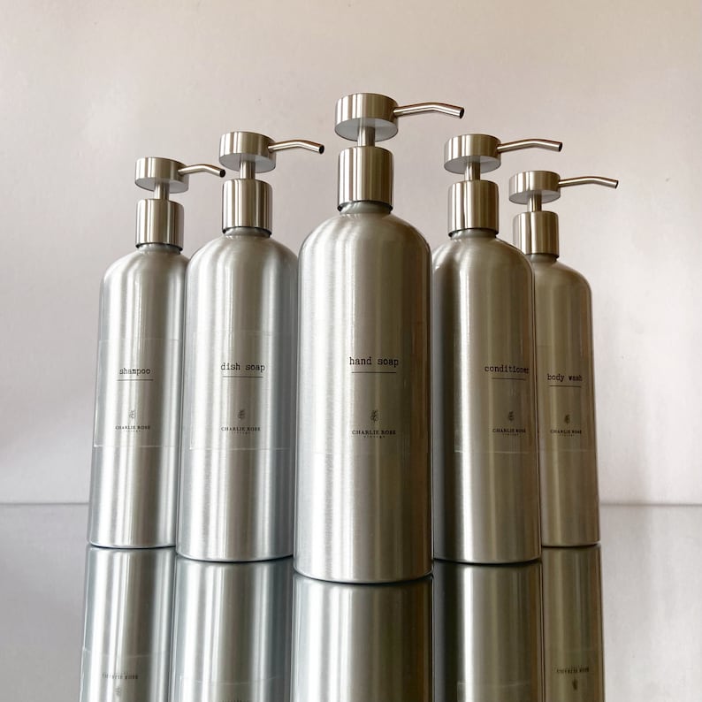 May include: Five silver pump bottles with labels for shampoo, dish soap, hand soap, conditioner, and body wash. The bottles are all the same size and shape, and they are arranged in a row on a white surface.