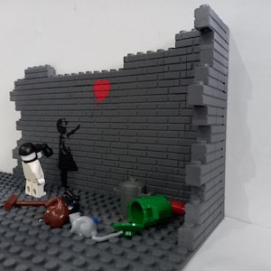 May include: A Lego model of a brick wall with a girl holding a red heart balloon. The girl is painted on the wall. There are other Lego pieces on the ground, including a trash can, a hammer, and a rat.
