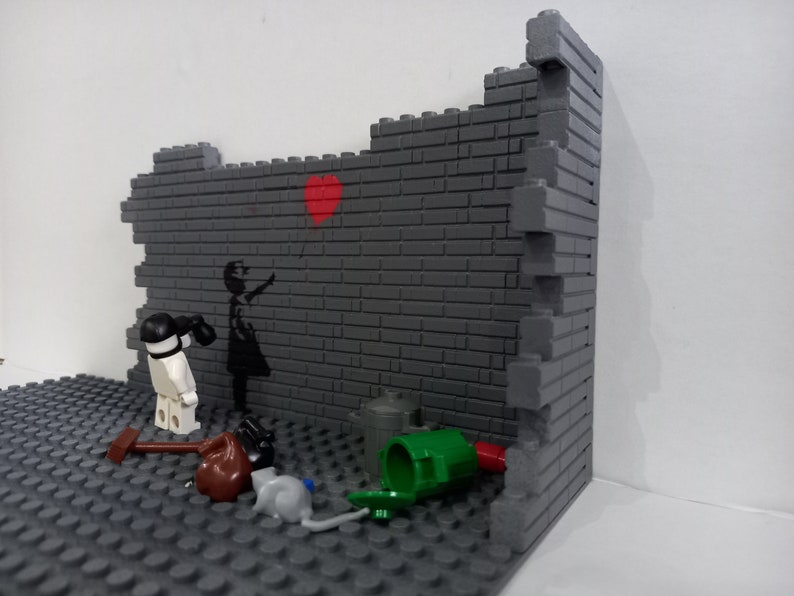May include: A Lego model of a brick wall with a girl holding a red heart balloon. The girl is painted on the wall. There are other Lego pieces on the ground, including a trash can, a hammer, and a rat.