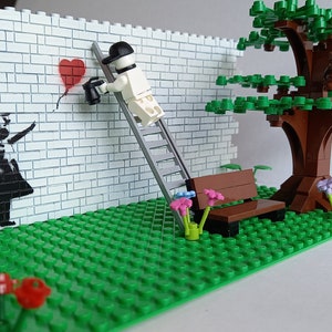 May include: A Lego model of a street scene with a brick wall, a lamppost, a bench, a tree, and a figure painting a red heart balloon on the wall. The figure is standing on a ladder.