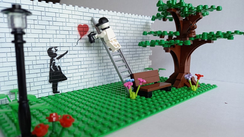 May include: A Lego model of a street scene with a brick wall, a lamppost, a bench, a tree, and a figure painting a red heart balloon on the wall. The figure is standing on a ladder.