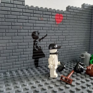 May include: A white Lego minifigure wearing a black cap and a black face mask stands in front of a gray brick wall. The wall has a black stencil of a girl holding a red heart balloon. The minifigure is holding a black broom and a black trash bag. There are other trash bags and a green trash can on the floor.