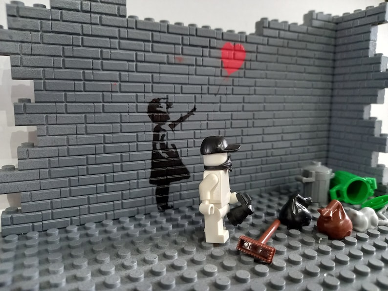 May include: A white Lego minifigure wearing a black cap and a black face mask stands in front of a gray brick wall. The wall has a black stencil of a girl holding a red heart balloon. The minifigure is holding a black broom and a black trash bag. There are other trash bags and a green trash can on the floor.