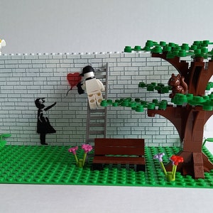 May include: A Lego model of a street scene with a white brick wall, a red heart balloon, a figure holding a paintbrush, a ladder, a bench, a tree with a squirrel, and a street lamp.