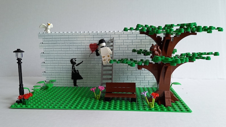 May include: A Lego model of a street scene with a white brick wall, a red heart balloon, a figure holding a paintbrush, a ladder, a bench, a tree with a squirrel, and a street lamp.
