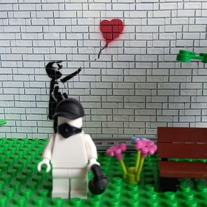 May include: A Lego minifigure wearing a black mask and cap stands in front of a brick wall with a black and white stencil of a child releasing a red heart-shaped balloon. The minifigure is standing on a green Lego baseplate with a brown Lego bench and green Lego plants.