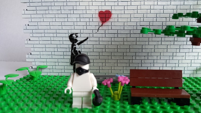 May include: A Lego minifigure wearing a black mask and cap stands in front of a brick wall with a black and white stencil of a child releasing a red heart-shaped balloon. The minifigure is standing on a green Lego baseplate with a brown Lego bench and green Lego plants.