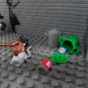 May include: A gray brick wall with a black and white stencil of a girl holding a balloon. There are several small toys on the floor in front of the wall, including a red bag, a gray rat, a green trash can, a red can, and a blue bag of chips.