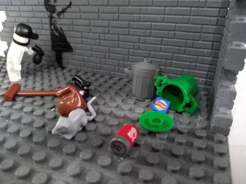 May include: A gray brick wall with a black and white stencil of a girl holding a balloon. There are several small toys on the floor in front of the wall, including a red bag, a gray rat, a green trash can, a red can, and a blue bag of chips.