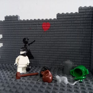 May include: A white Lego figure wearing a black helmet and mask stands in front of a gray brick wall with a black and white stencil of a girl holding a red heart balloon. There are two trash cans, a brown bag, and a gray rat on the ground.