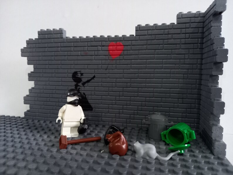 May include: A white Lego figure wearing a black helmet and mask stands in front of a gray brick wall with a black and white stencil of a girl holding a red heart balloon. There are two trash cans, a brown bag, and a gray rat on the ground.