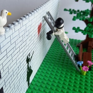 May include: A Lego scene with a white brick wall, a red heart painted on the wall, a figure on a ladder painting the heart, a green lawn, a tree with a brown bench, and a white seagull on top of the wall.
