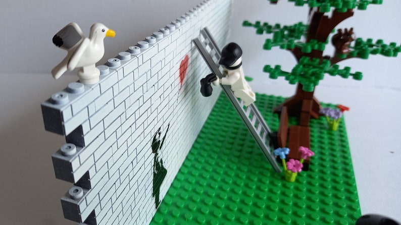 May include: A Lego scene with a white brick wall, a red heart painted on the wall, a figure on a ladder painting the heart, a green lawn, a tree with a brown bench, and a white seagull on top of the wall.