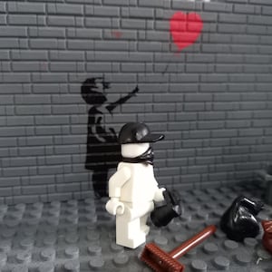 May include: A white Lego figure wearing a black cap and bandana stands in front of a brick wall with a black and white stencil of a child holding a red heart balloon. The figure is holding a black trash bag.
