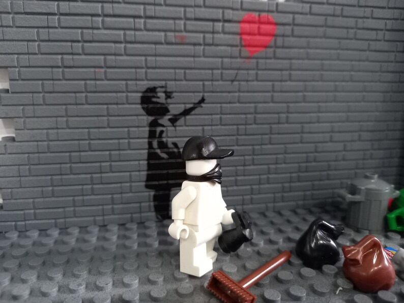May include: A white Lego figure wearing a black cap and bandana stands in front of a brick wall with a black and white stencil of a child holding a red heart balloon. The figure is holding a black trash bag.
