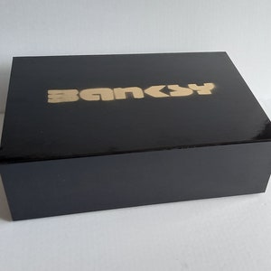 May include: A black box with the word "Banksy" printed in gold letters on the top.