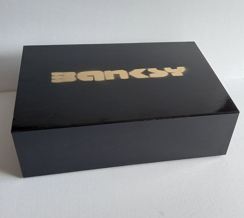 May include: A black box with the word "Banksy" printed in gold letters on the top.