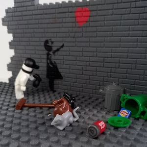 May include: A Lego scene depicting a brick wall with a black and white stencil of a girl holding a red heart balloon. A Lego figure of a person in a white shirt and black hat is standing in front of the wall. There are other Lego pieces scattered on the ground, including a brown bucket, a green trash can, a gray rat, and a red can with the word "Crisps" on it.