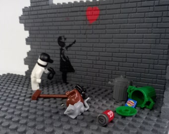 May include: A Lego scene featuring a brick wall with a black and white stencil of a girl holding a red heart balloon. A Lego figure of a person in a white shirt and black hat is standing in front of the wall. There are other Lego pieces scattered on the ground, including a brown bucket, a green bin, a grey rat, and a red can with the word "Crisps" on it.
