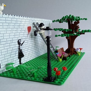 May include: A Lego model of a street scene with a brick wall, a girl holding a red balloon, a man on a ladder painting a heart, a tree, a bench, and a street lamp.