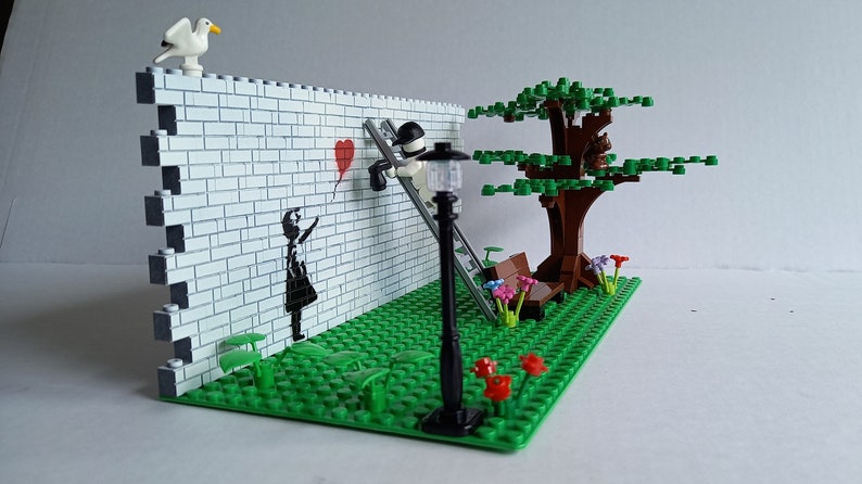May include: A Lego model of a street scene with a brick wall, a girl holding a red balloon, a man on a ladder painting a heart, a tree, a bench, and a street lamp.