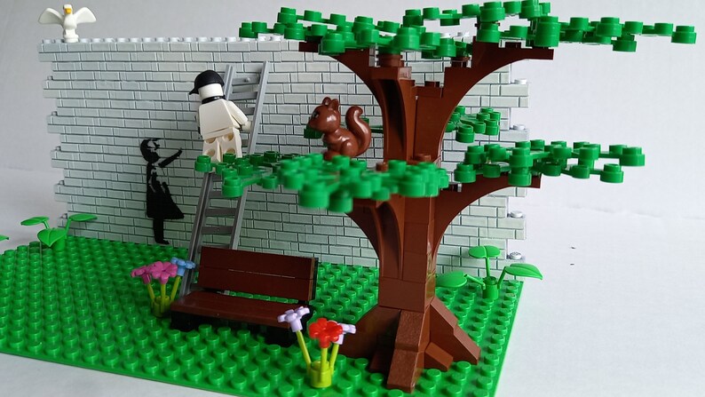 May include: A Lego model of a park scene with a brick wall, a tree, a bench, and a figure painting a mural. The mural depicts a girl holding a balloon. A squirrel sits on a branch of the tree.