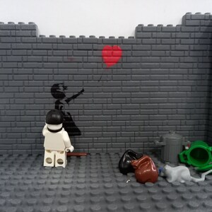May include: A Lego minifigure stands in front of a brick wall with a black and white stencil of a girl letting go of a red balloon. The wall also has a red heart painted on it. There are several other Lego pieces on the floor, including a green trash can, a brown trash bag, and a gray cat.