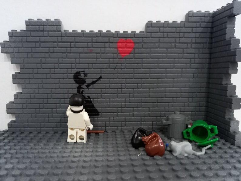 May include: A Lego minifigure stands in front of a brick wall with a black and white stencil of a girl letting go of a red balloon. The wall also has a red heart painted on it. There are several other Lego pieces on the floor, including a green trash can, a brown trash bag, and a gray cat.