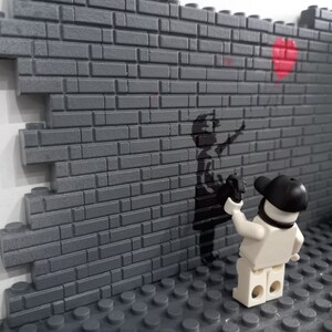 May include: A white Lego figure with a black cap is spray painting a black stencil of a child holding a red balloon on a gray brick wall.