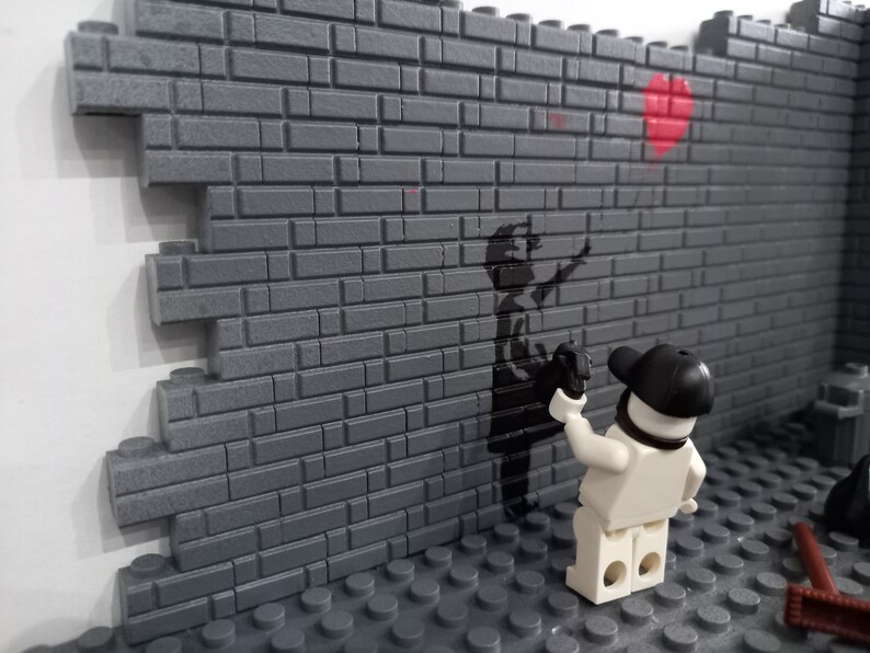 May include: A white Lego figure with a black cap is spray painting a black stencil of a child holding a red balloon on a gray brick wall.
