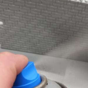May include: A hand holding a blue and silver spray can, spraying a mist onto a gray brick wall.