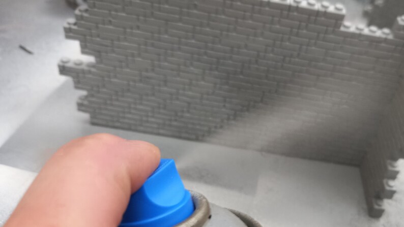 May include: A hand holding a blue and silver spray can, spraying a mist onto a gray brick wall.