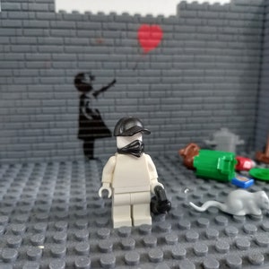 May include: A white Lego minifigure wearing a black cap and bandana stands on a gray Lego baseplate. The minifigure is holding a black gun. The background is a gray brick wall with a black stencil of a girl holding a balloon.