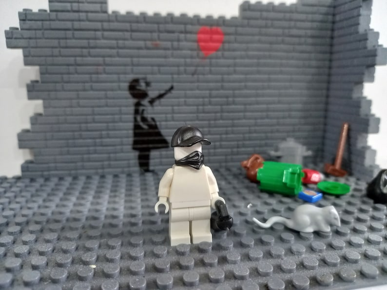 May include: A white Lego minifigure wearing a black cap and bandana stands on a gray Lego baseplate. The minifigure is holding a black gun. The background is a gray brick wall with a black stencil of a girl holding a balloon.