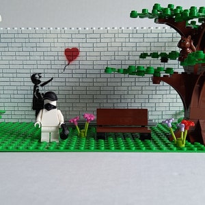 May include: A Lego model of a park scene with a white brick wall, a red heart balloon, a black and white figure, a white figure with a black mask, a brown bench, a green tree with a brown trunk, and green grass.