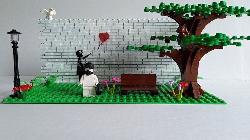 May include: A Lego model of a park scene with a white brick wall, a red heart balloon, a black and white figure, a white figure with a black mask, a brown bench, a green tree with a brown trunk, and green grass.