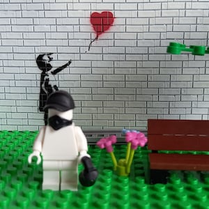 May include: A white Lego figure wearing a black mask and hat stands in front of a brick wall with a black and white stencil of a child reaching for a red heart-shaped balloon. The figure is holding a black object in its right hand. There is a brown wooden bench and green plants in the foreground.