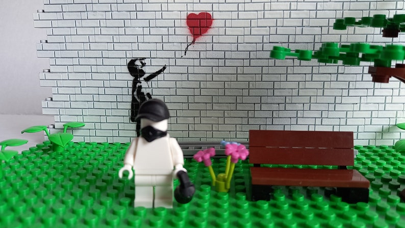 May include: A white Lego figure wearing a black mask and hat stands in front of a brick wall with a black and white stencil of a child reaching for a red heart-shaped balloon. The figure is holding a black object in its right hand. There is a brown wooden bench and green plants in the foreground.